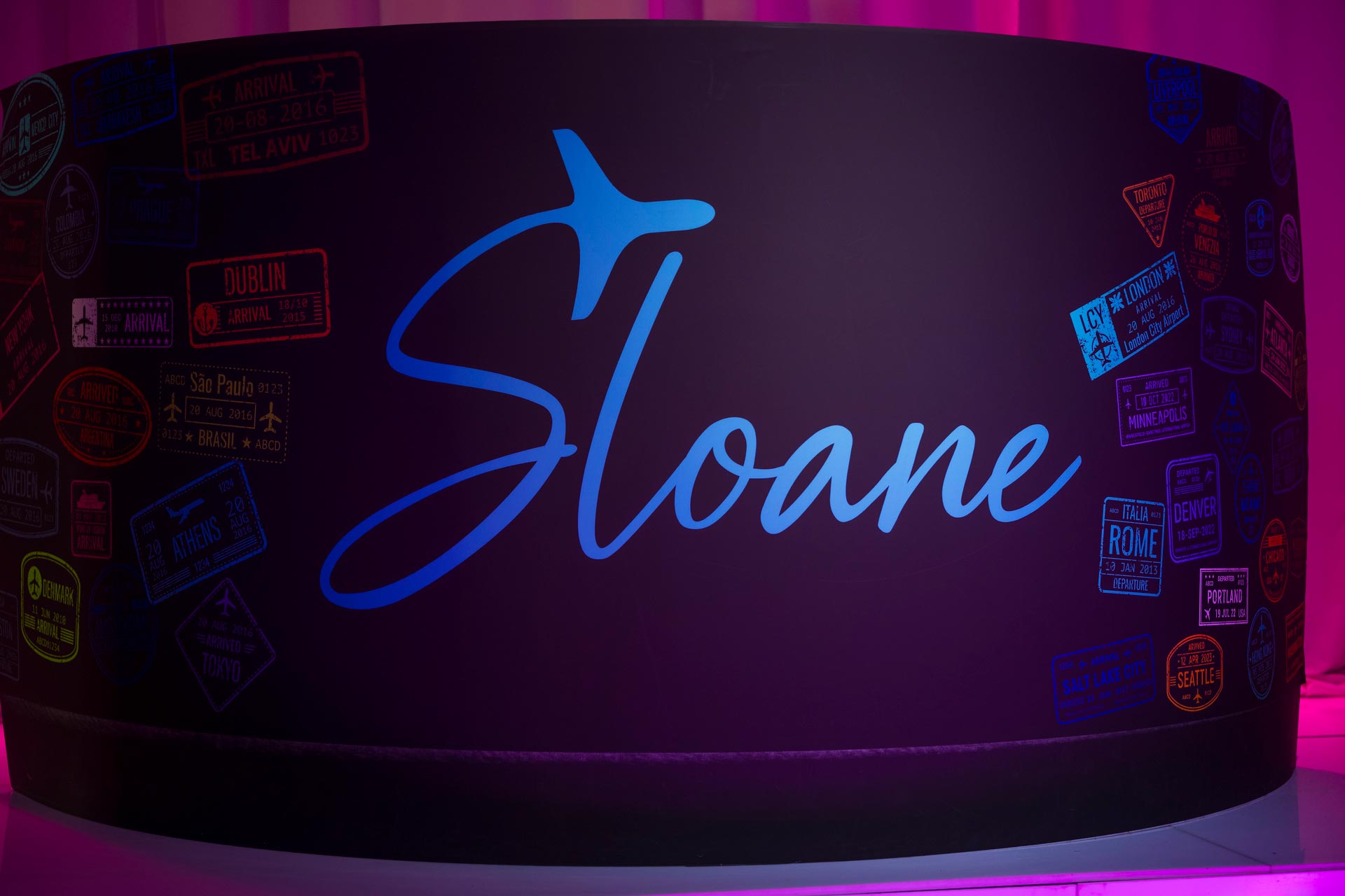 The image shows a sign with the name  Sloane  on it, surrounded by various stickers and logos, placed against a dark background with colorful lighting.